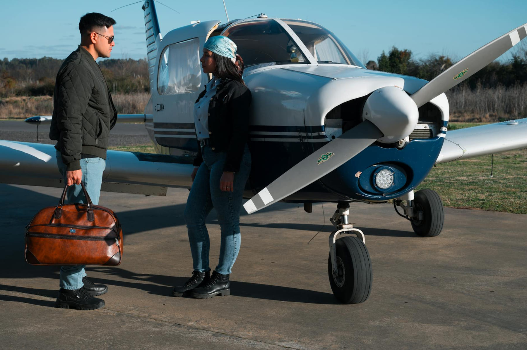 Young couple standing by a private airplane, conversing and holding a bag, at a sunny airfield. on Miles and Memories