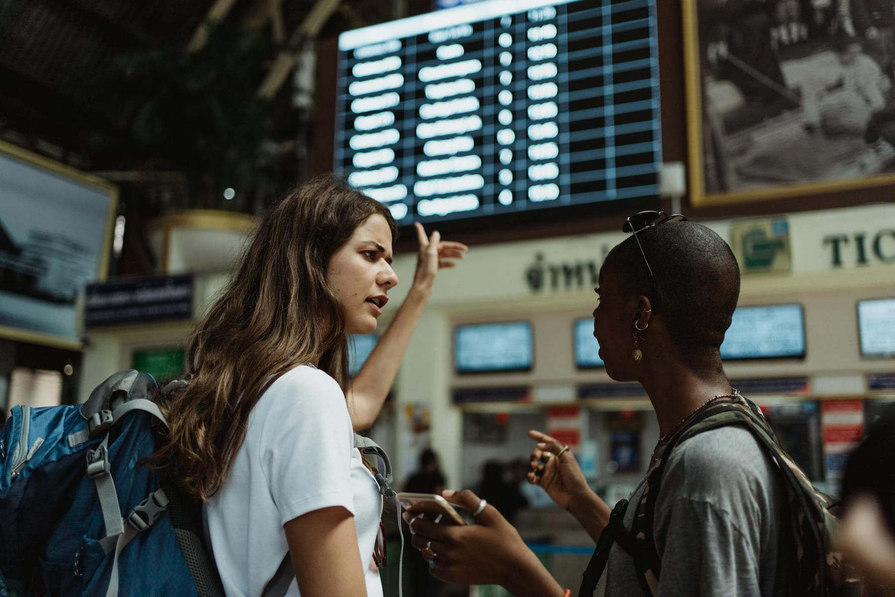 Two travelers with backpacks discuss in front of an airport timetable. Adventure awaits. on Miles and Memories